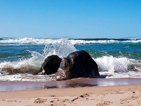 A wave breaks against a boulder embedded in the sand